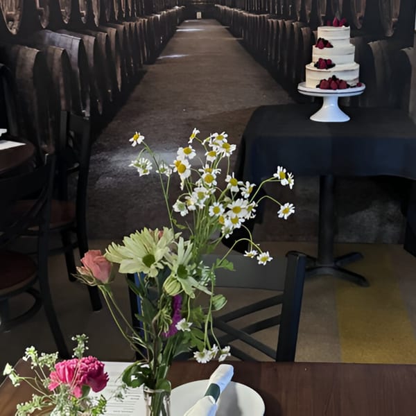 A three-tiered cake with berries on a table, with flowers in the foreground and a barrel-lined hallway behind.