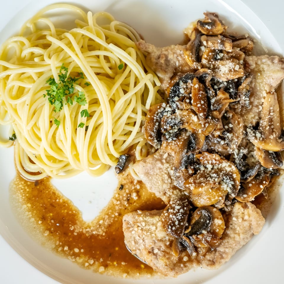 Veal scallopini with mushroom sauce and spaghetti, garnished with parsley, on a white plate.